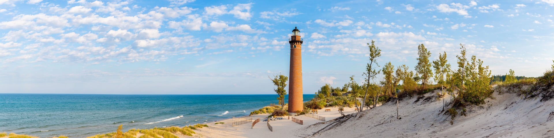A coastal landscape with a lighthouse, sandy beach, grassy dunes, and a clear blue sky.