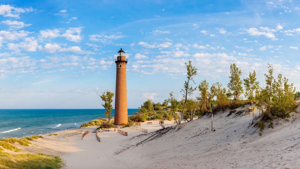 A coastal landscape with a lighthouse, sandy beach, grassy dunes, and a clear blue sky.
