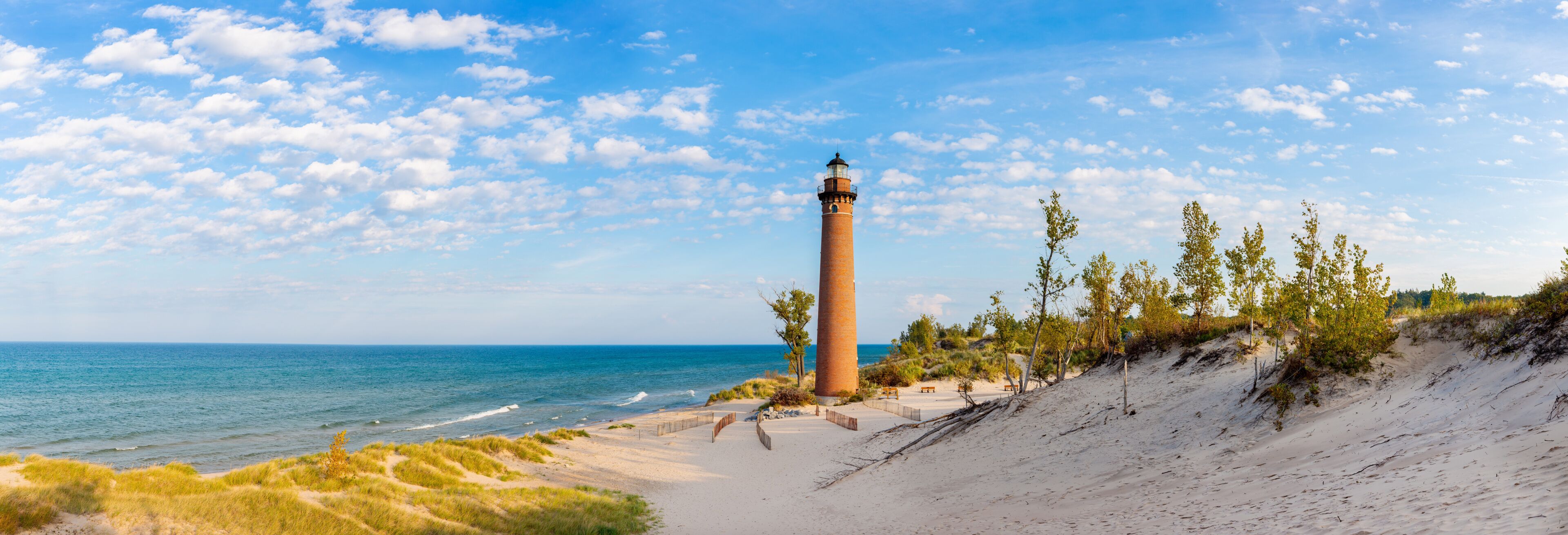 A coastal landscape with a lighthouse, sandy beach, grassy dunes, and a clear blue sky.