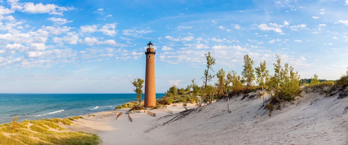 A coastal landscape with a lighthouse, sandy beach, grassy dunes, and a clear blue sky.