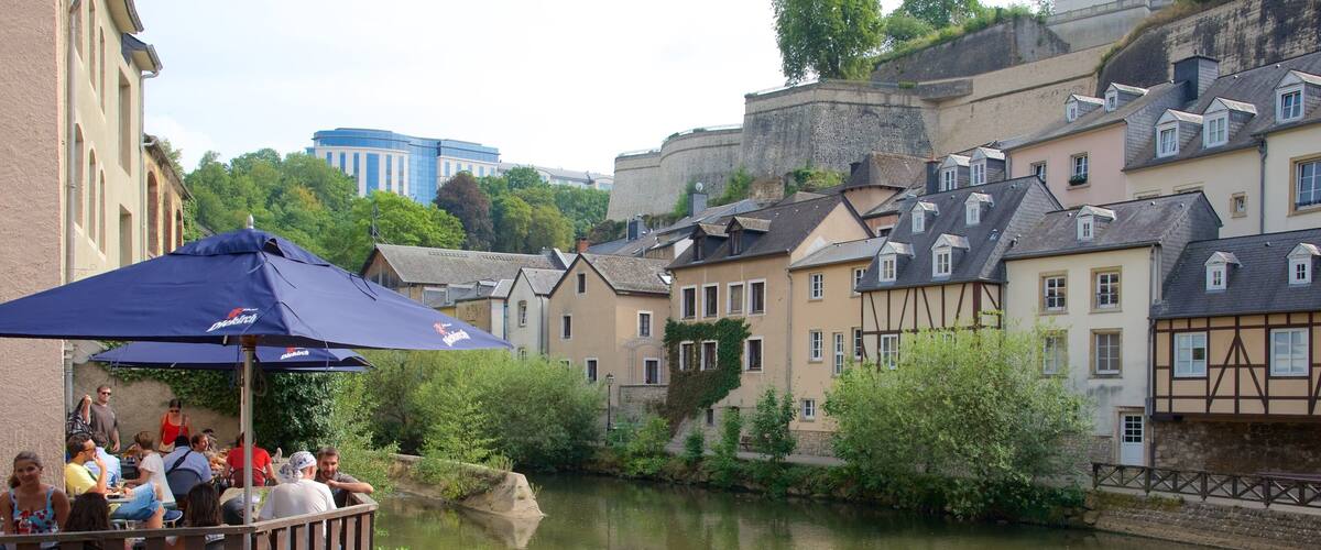 A riverside scene with people dining under umbrellas, traditional buildings, and a castle in the background.