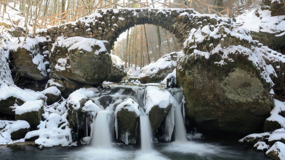 A stone bridge over a flowing river with snow-covered rocks and trees in the background.