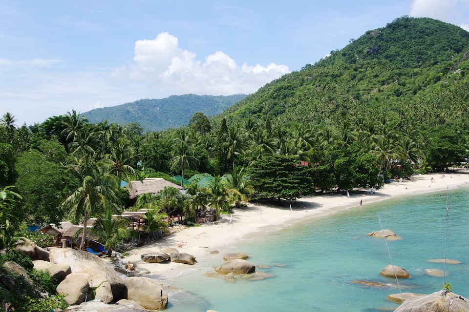Uma praia tropical com águas turquesa cristalinas, areia branca e colinas verdes exuberantes ao fundo.