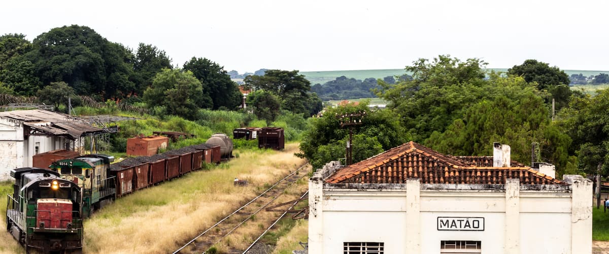 A train on tracks, a white building labeled "MATAO," and lush greenery in the background.
