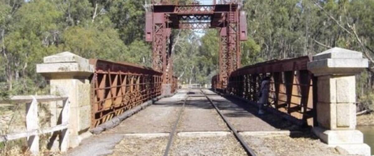 A railway bridge with a metal arch structure, surrounded by trees and a clear sky.
