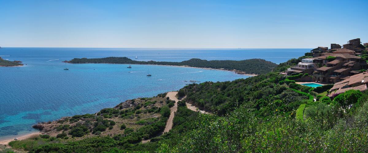A coastal landscape with clear blue waters, a sandy beach, and a cluster of traditional houses on a hillside.
