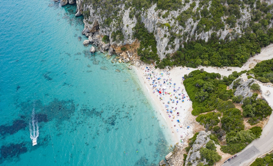 View from above, stunning aerial view of a beautiful beach full of beach umbrellas and people sunbathing and swimming on a turquoise water. Cala Gonone, Sardinia, Italy.