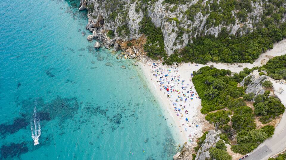 A coastal beach with clear turquoise waters, rocky cliffs, and dense greenery.