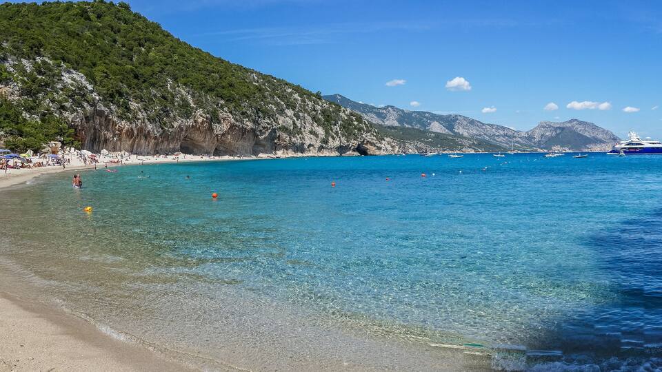 A beach with clear turquoise water, a rocky cliff, and boats in the distance.