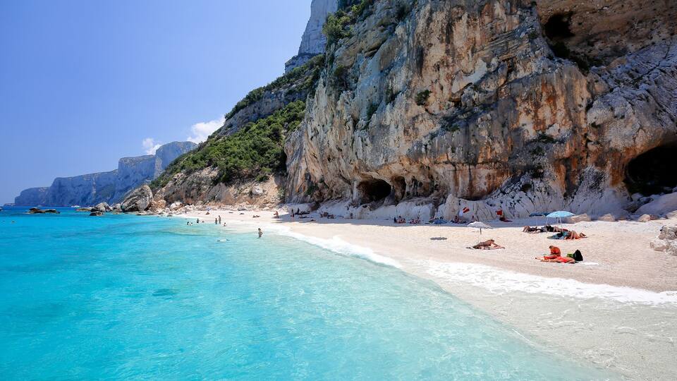 A beach with clear turquoise water, white sand, and rocky cliffs with caves.