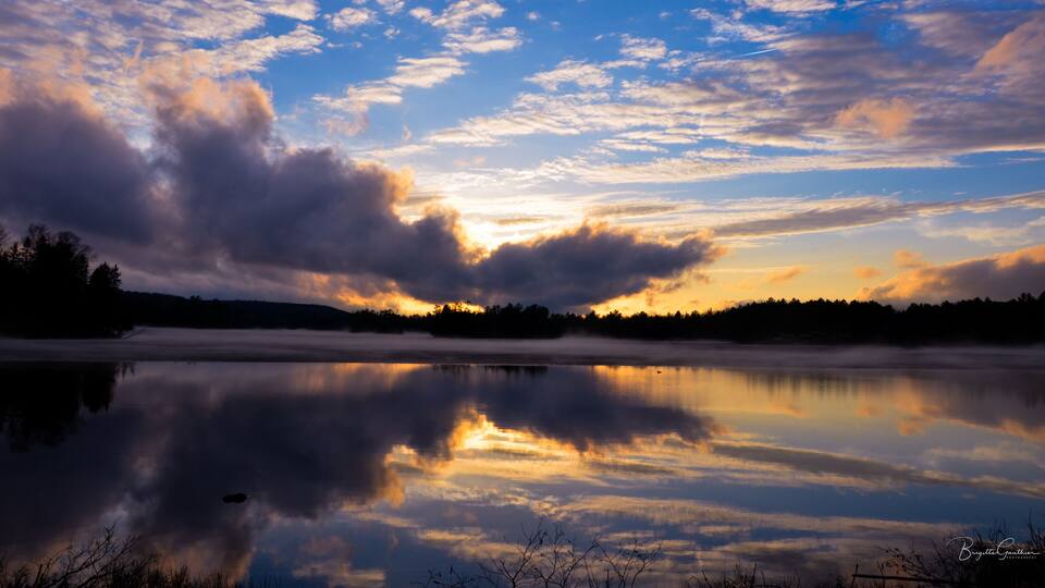 A calm lake reflecting the sky with clouds and a forest in the background.