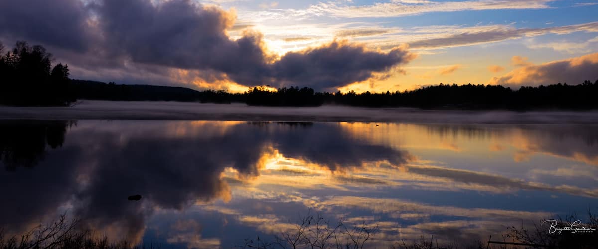 A calm lake reflecting the sky with clouds and a forest in the background.