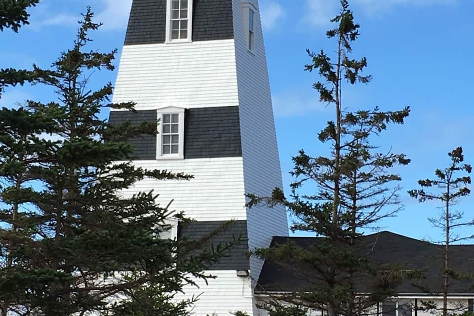 Un grand phare, coiffé d’un dôme rouge, entouré d’arbres et d’une maison.