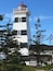 Un grand phare, coiffé d’un dôme rouge, entouré d’arbres et d’une maison.
