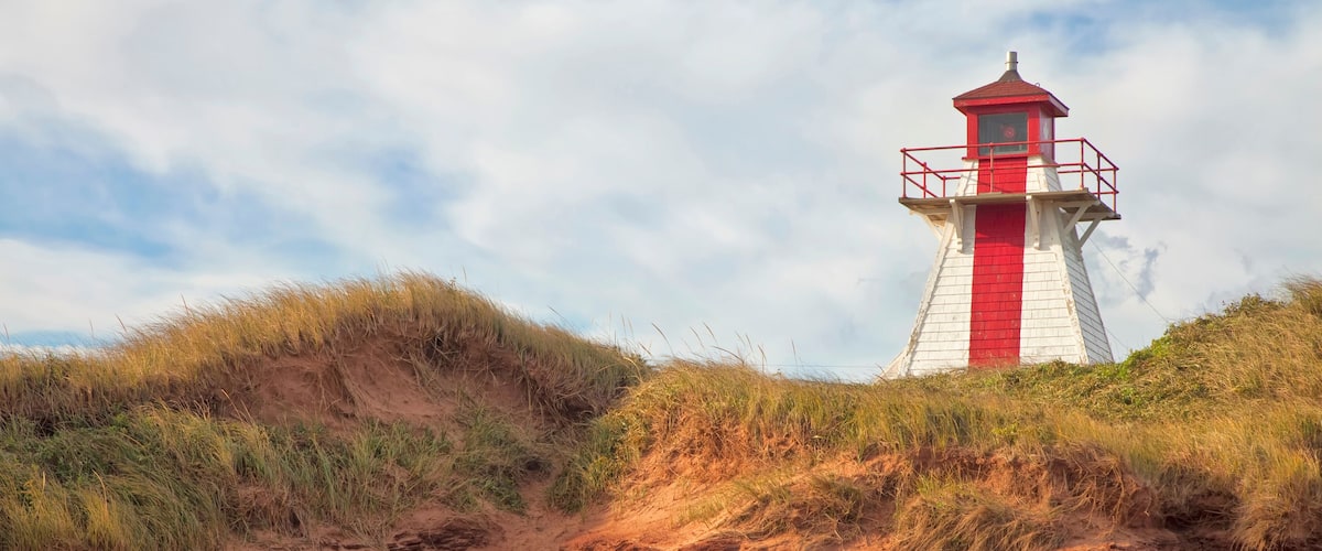 A red and white lighthouse on a sandy beach with dunes and rocky outcrops.