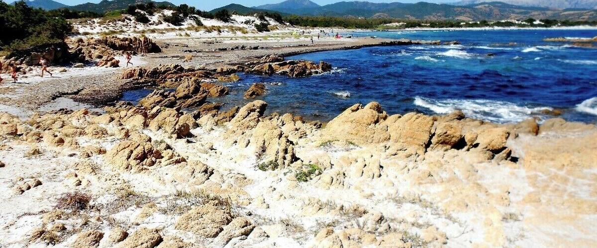 A rocky shoreline with waves crashing onto the beach, a clear blue sky, and distant mountains.