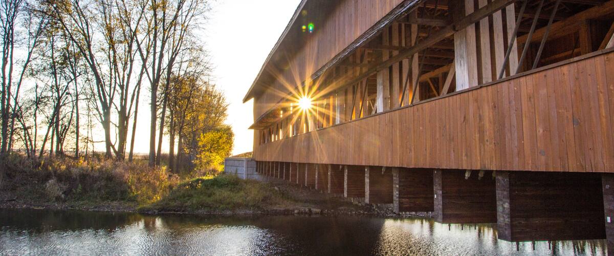A wooden covered bridge over a calm river, surrounded by trees and a clear sky.