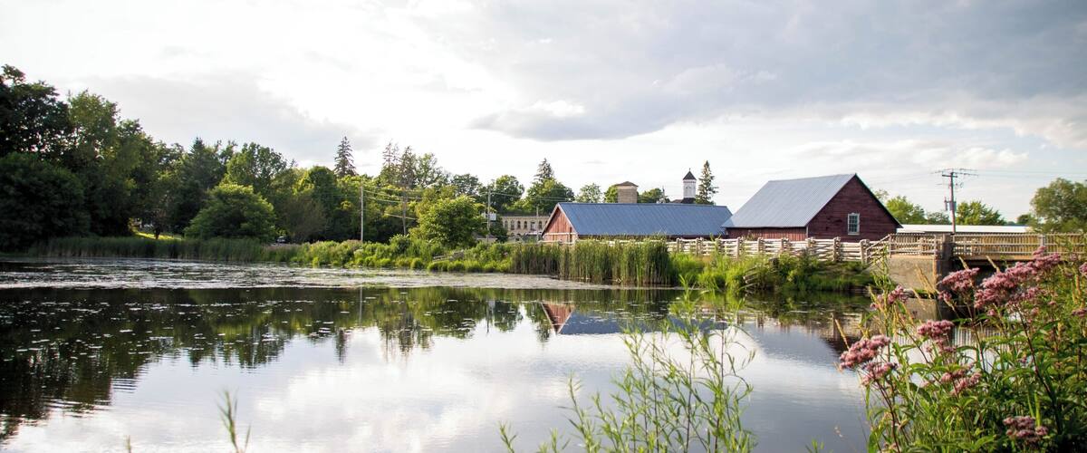 A calm river reflecting a red barn and a white church, surrounded by lush greenery and trees.