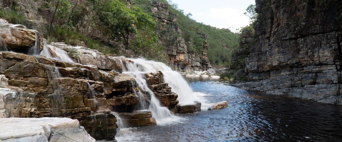 A waterfall cascading over rocky terrain into a river, surrounded by dense forest and steep cliffs.