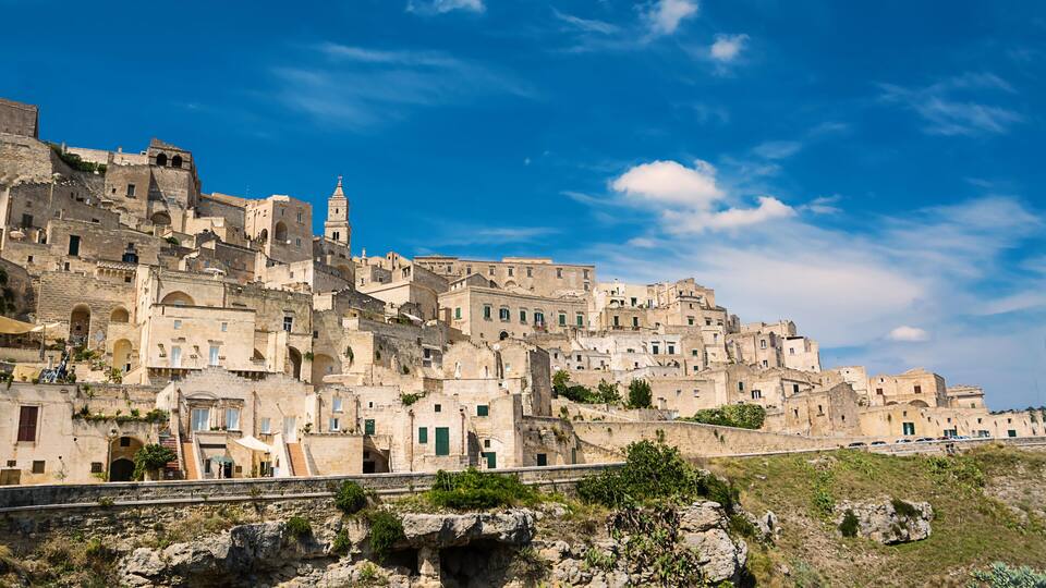 A historic town with ancient stone buildings, a prominent church tower, and a rocky hillside.