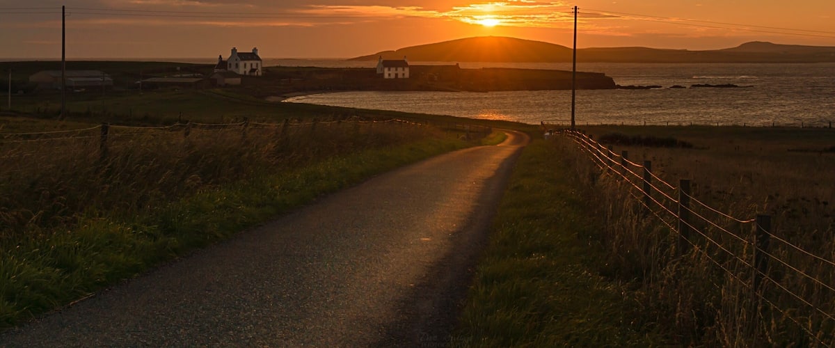 A coastal road leading to a small island with houses, surrounded by grass and a fence.