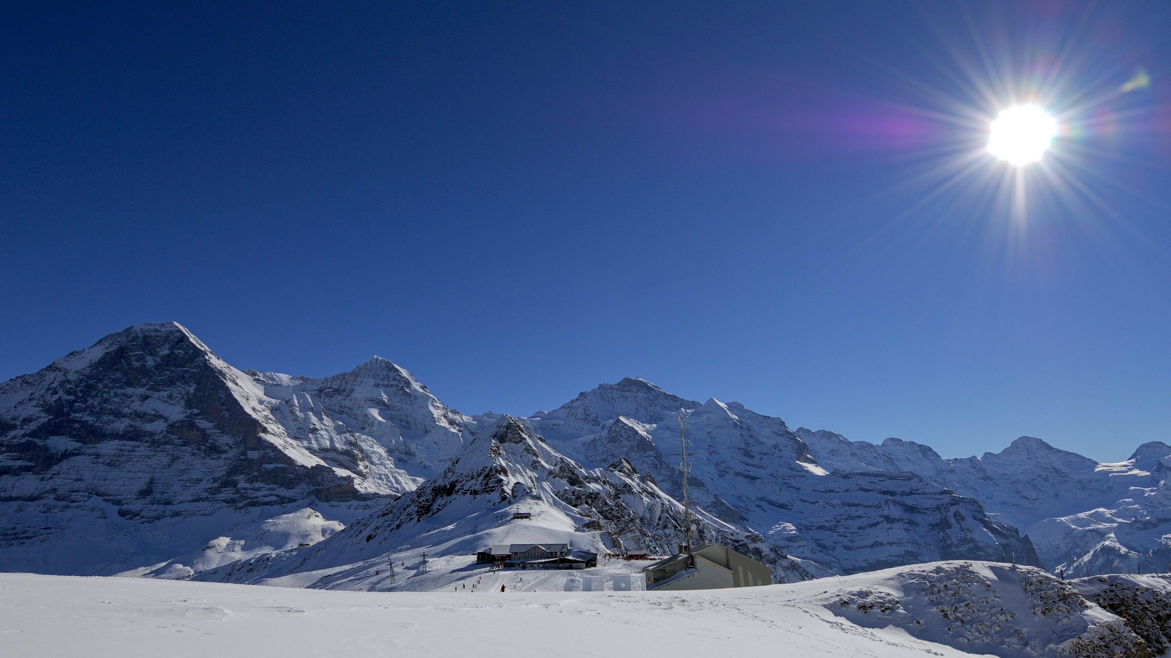 A snow-covered mountain range with a bright sun in the sky and a lodge in the foreground.