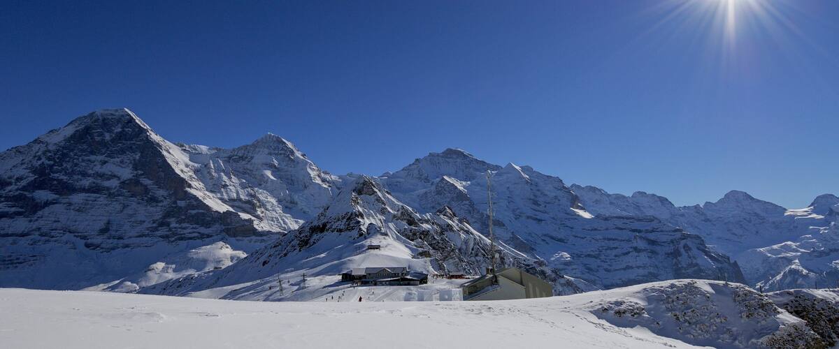 A snow-covered mountain range with a bright sun in the sky and a lodge in the foreground.