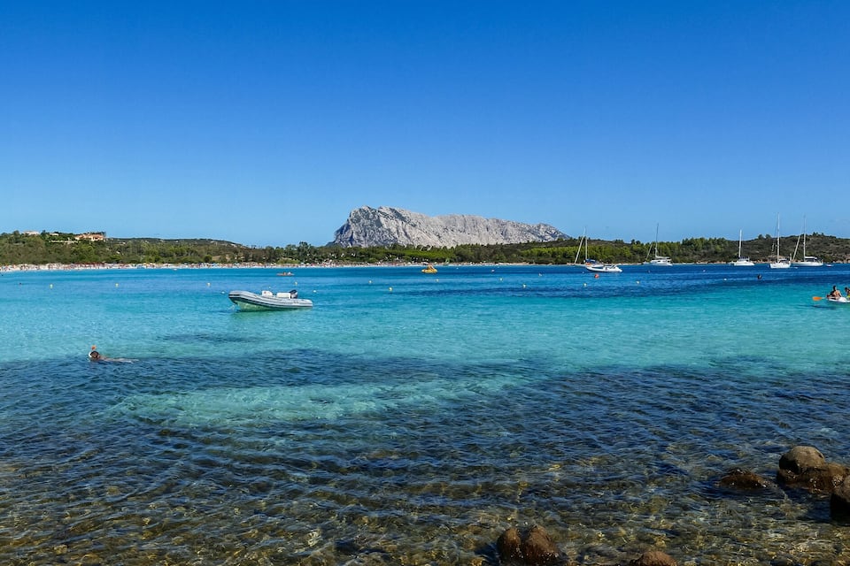 A clear blue sea with boats, a rocky shore, and a mountain in the background.