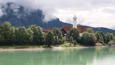 Un pueblo junto al lago con una torre de iglesia, rodeado de exuberante vegetación y montañas al fondo.