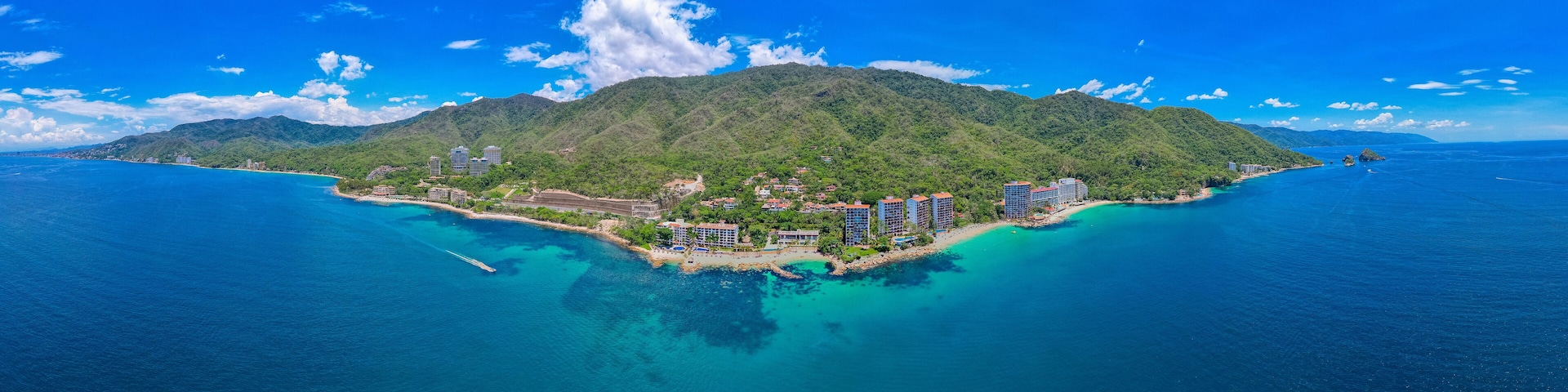 A tropical island with a clear blue sea, lush green mountains, and several high-rise buildings.
