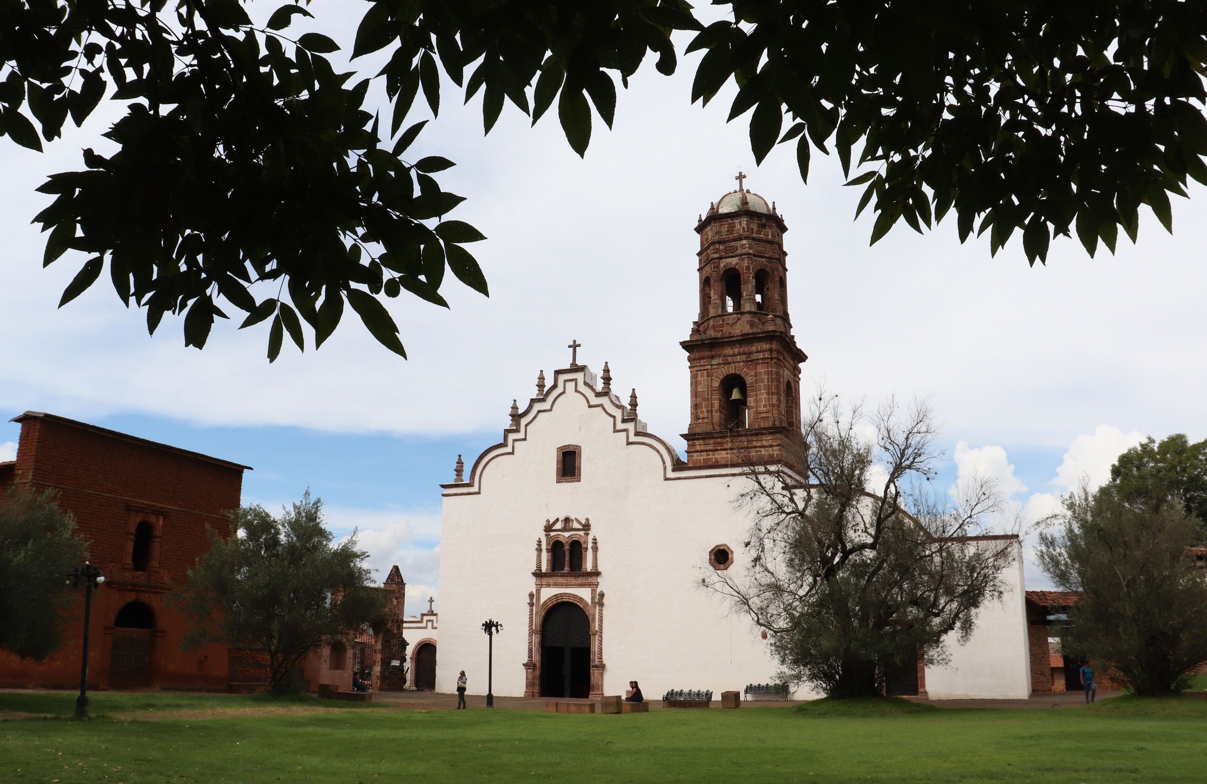 A white church with a bell tower and arched windows, surrounded by trees and a grassy field.