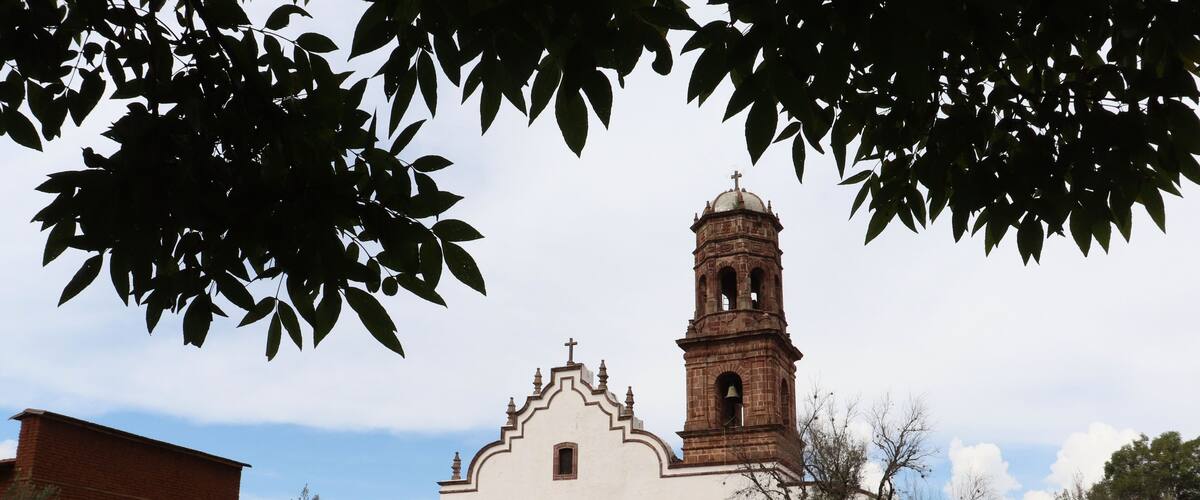 A white church with a bell tower and arched windows, surrounded by trees and a grassy field.