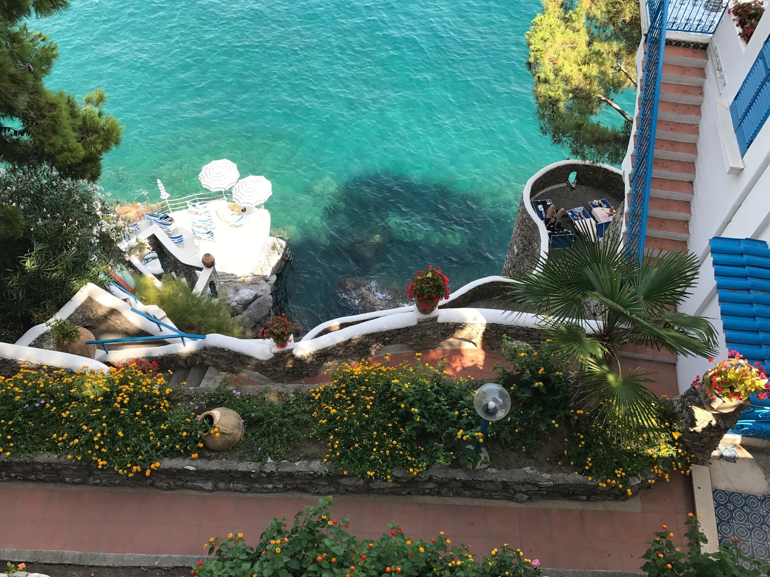 A coastal view with clear turquoise water, a stone pathway, and a white building with blue shutters.