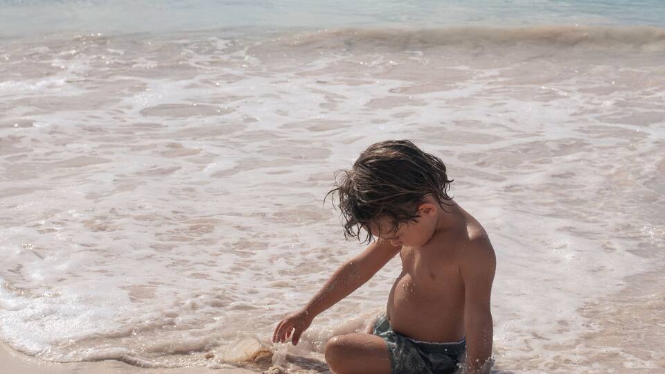A child playing in the shallow water at the beach with boats in the background.