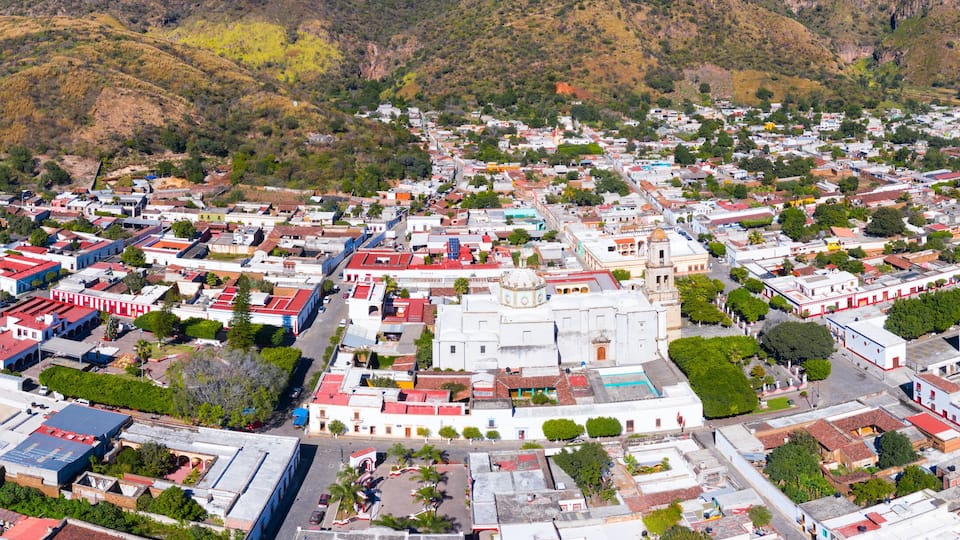 An aerial view of a town with a central plaza, surrounded by residential and commercial buildings, with hills in the background.