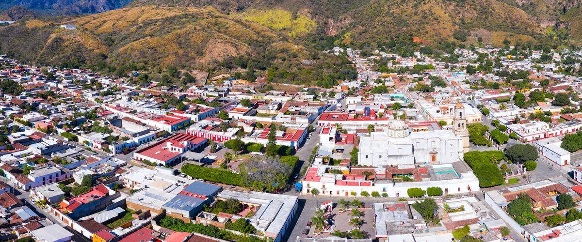 An aerial view of a town with a central plaza, surrounded by residential and commercial buildings, with hills in the background.