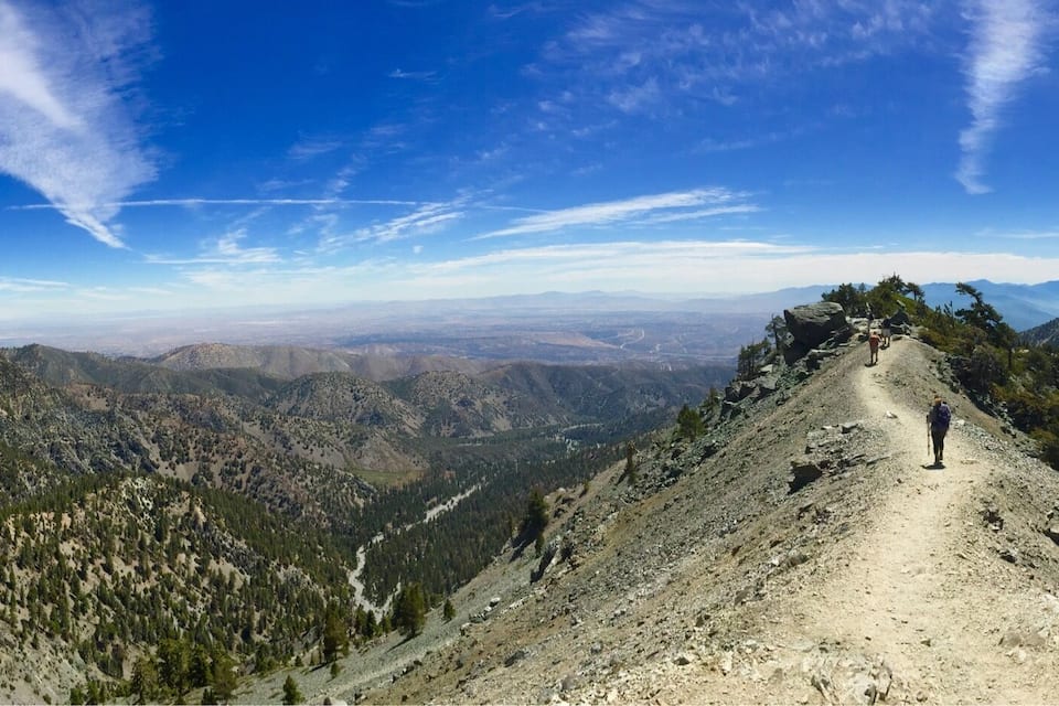 A mountain trail with a person hiking, surrounded by forested slopes and distant mountain ranges under a clear blue sky.