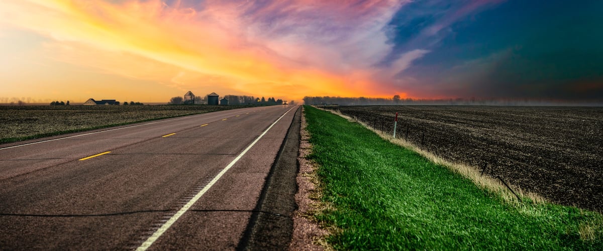 A rural road with fields on both sides, a farmhouse, and a barn in the distance under a colorful sky.
