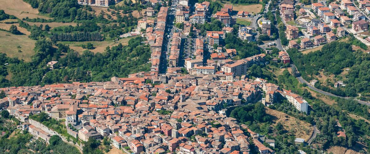 An aerial view of a densely packed town with red-roofed buildings, surrounded by greenery and hilly terrain.