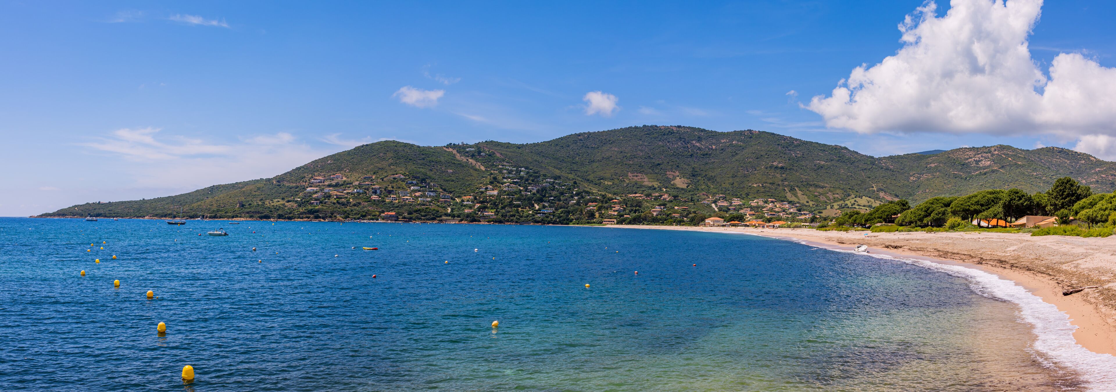 A coastal landscape with a clear blue sea, a sandy beach, and a hill with buildings in the background.