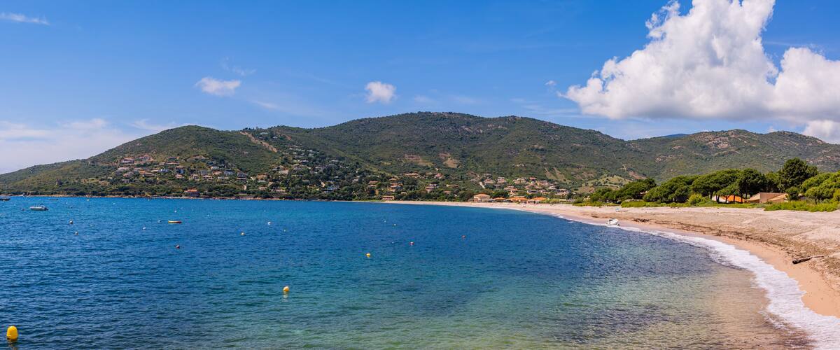 A coastal landscape with a clear blue sea, a sandy beach, and a hill with buildings in the background.