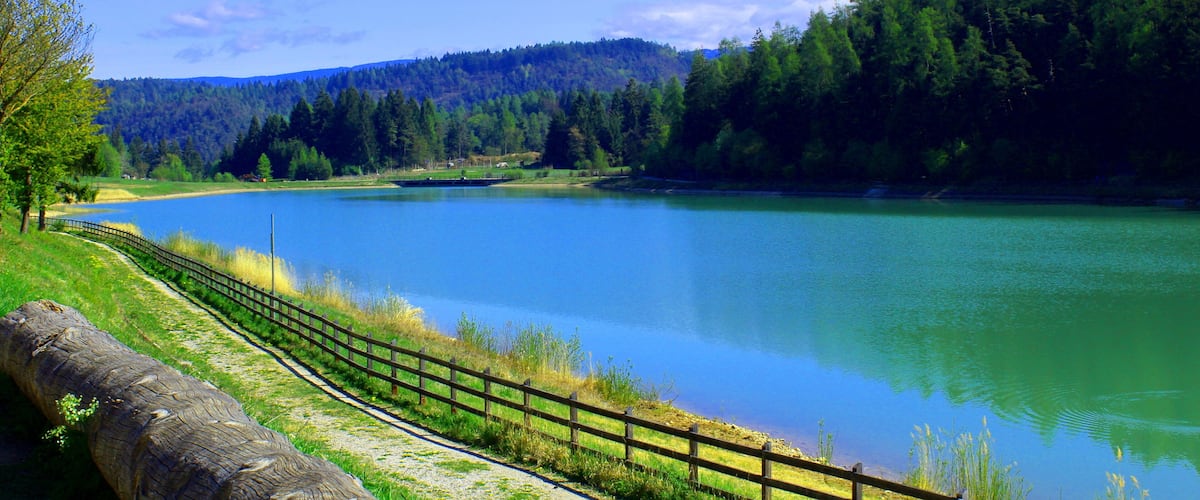 A calm lake surrounded by lush green hills and a wooden fence along the shore.