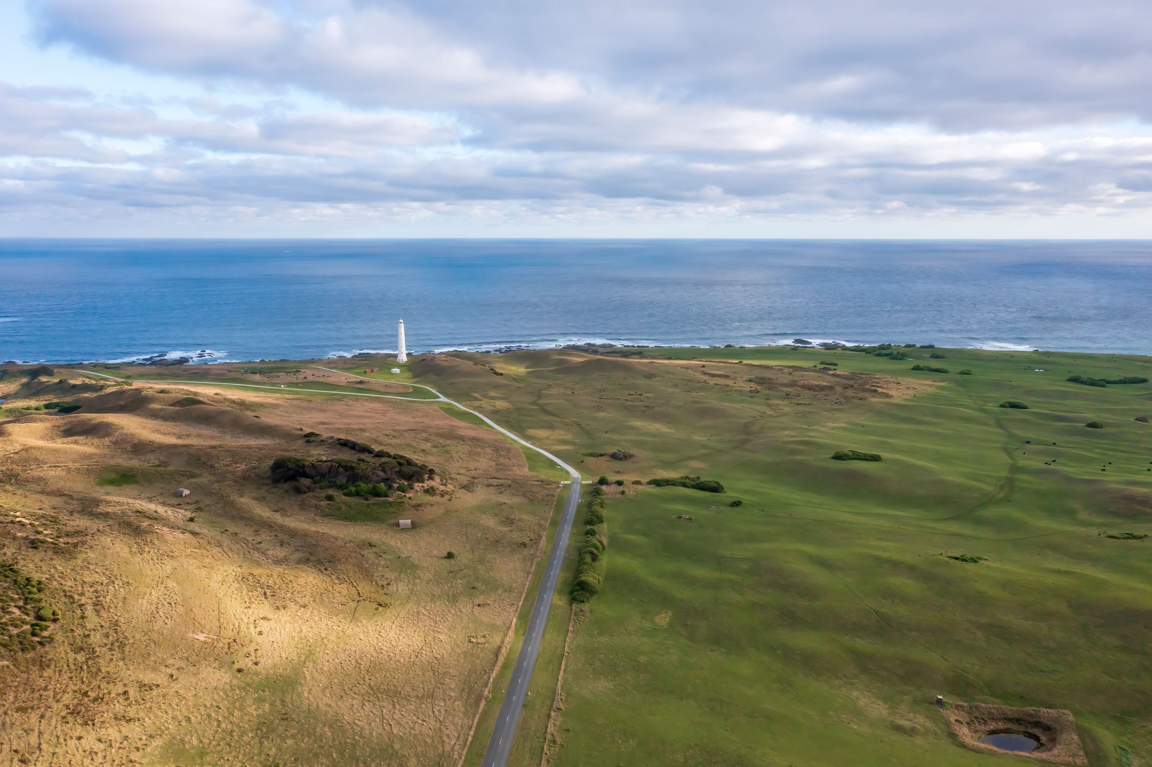 A coastal landscape with a lighthouse, a road, and a grassy field.