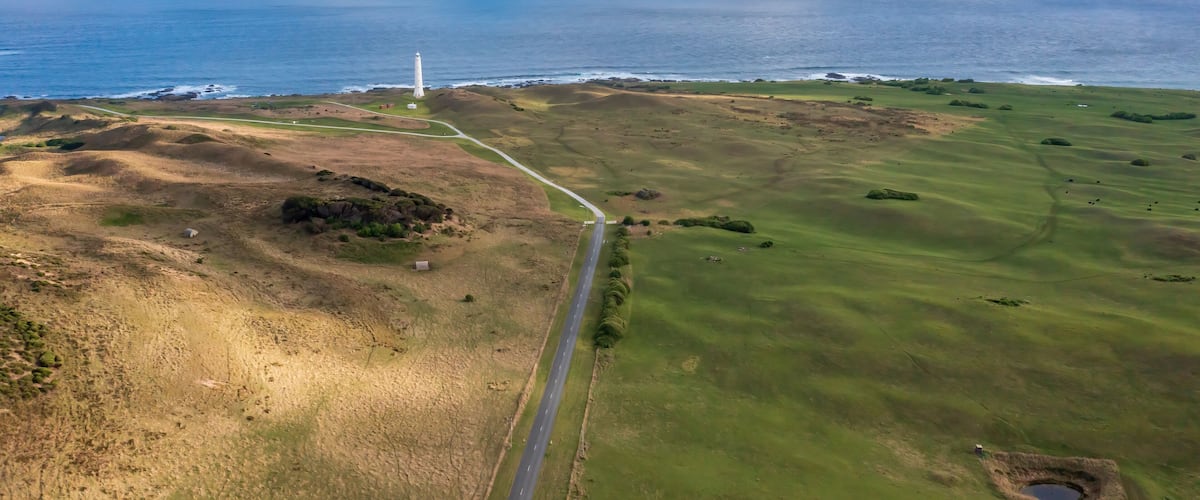 A coastal landscape with a lighthouse, a road, and a grassy field.
