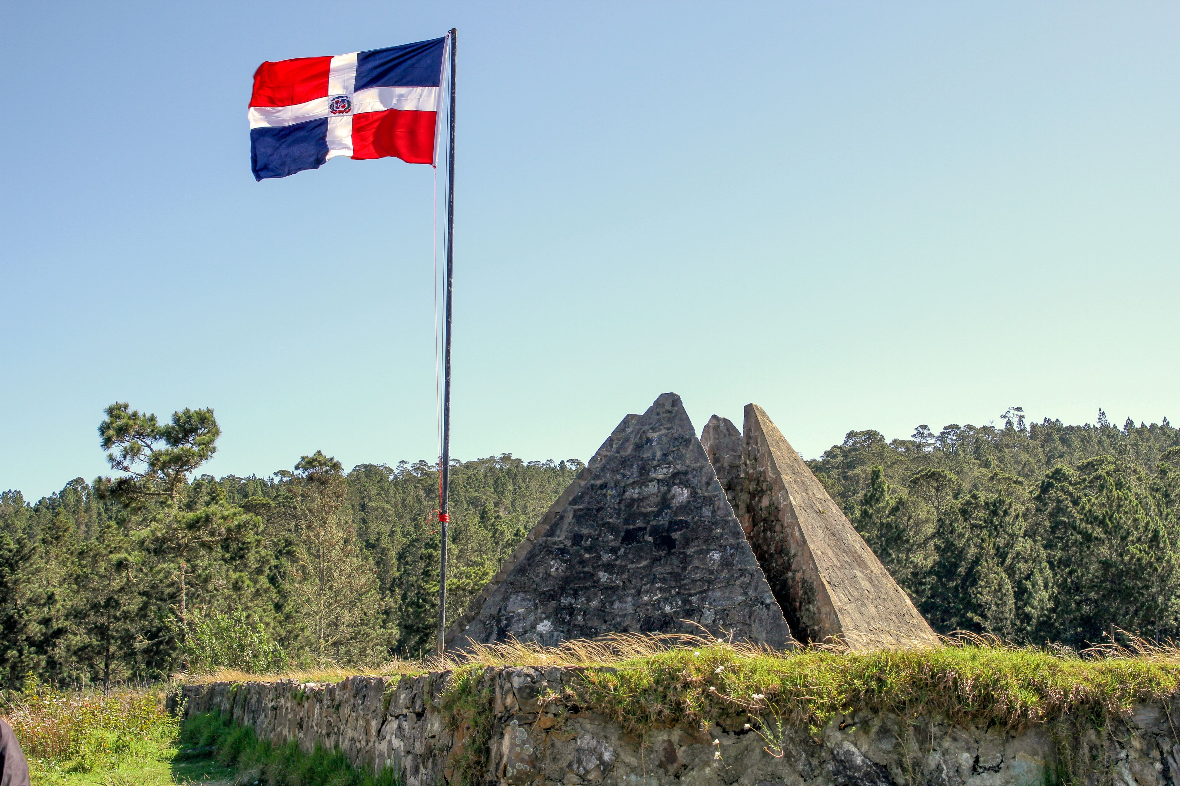 Um mastro com uma bandeira, duas estruturas de pedra piramidais e um fundo arborizado.