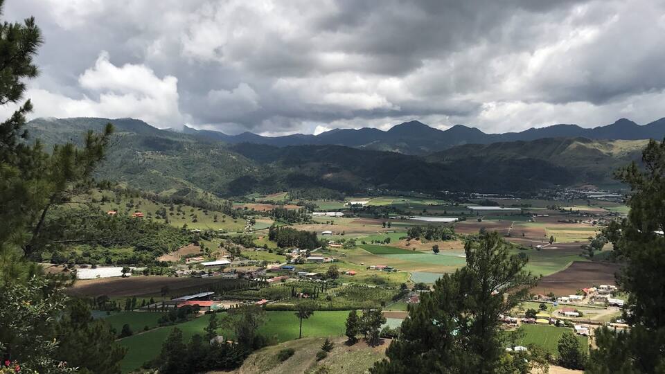 A mountainous landscape with fields, a small village, and distant mountains under a cloudy sky.