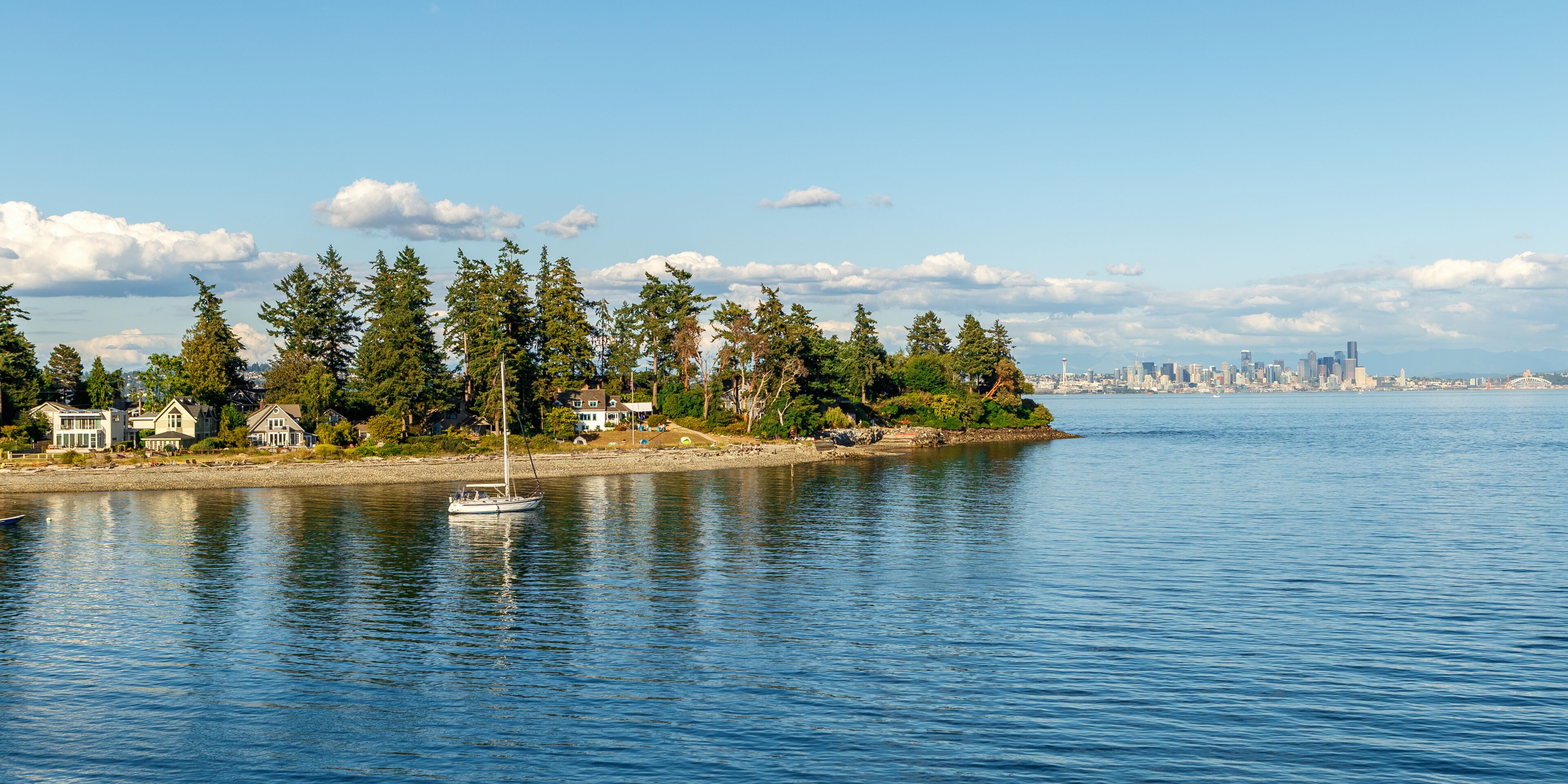 A calm waterfront with a sailboat, houses, trees, and a distant city skyline.