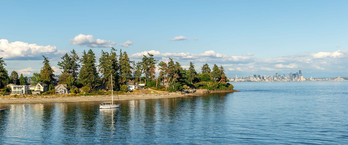 A calm waterfront with a sailboat, houses, trees, and a distant city skyline.