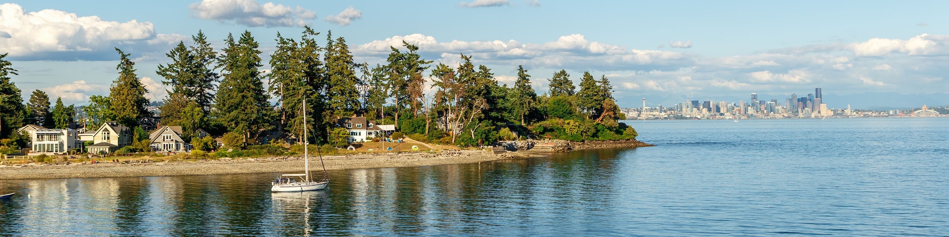 A calm waterfront with a sailboat, houses, trees, and a distant city skyline.