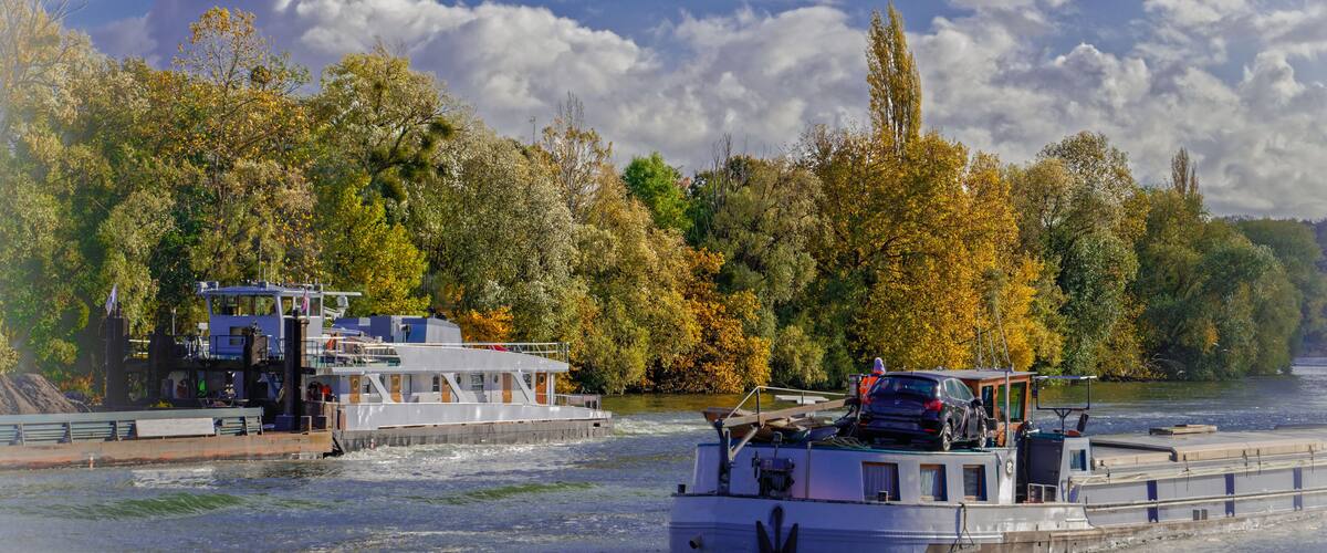 A river with two boats, one docked and one moving, surrounded by autumn-colored trees.